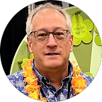 A man wearing glasses, a blue patterned shirt, and an orange lei stands in front of a green display with clover-shaped signs.